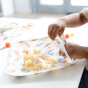 A child plays with pasta and pom-poms for sensory fun using Bumkins Clear Travel Bag 3-Pack: Winnie and Friends at a white table.