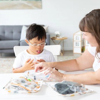 A child with glasses and an adult explore Bumkins Clear Travel Bag 3-Pack: Winnie and Friends on a white table in a cozy living room.