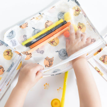 Close-up of a child's hands arranging colorful markers in a Bumkins Clear Travel Bag 3-Pack: Winnie and Friends on a white table.