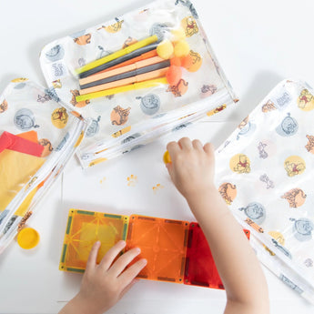 A child plays with magnetic tiles near Bumkins Clear Travel Bag 3-Pack: Winnie and Friends filled with pom-poms and markers on a table.