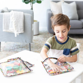 A child plays with markers and art supplies in Bumkins Clear Travel Bag 3-Pack: Super Mario™ Mashup at a white table in a cozy living room.