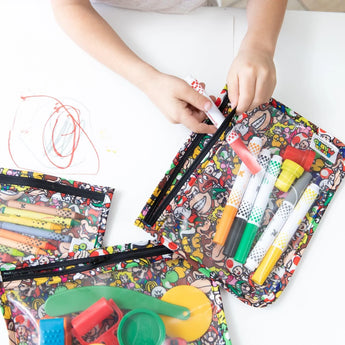 On a white table, a child organizes markers and art supplies into Bumkins Clear Travel Bag 3-Pack: Super Mario™ Mashup.