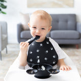 In a high chair, a toddler smiles in a Bumkins Silicone Bib: Mickey Mouse Faces, holding a spoon with a bowl in front.