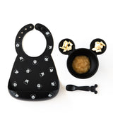 Bumkins Silicone Bib: Mickey Mouse Faces, sits next to a bowl with applesauce & banana chunks and a matching spoon on a white background.
