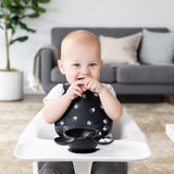 A baby in a high chair wears a Bumkins Silicone Bib: Mickey Mouse Faces, enjoying snacks from a black bowl. A grey sofa and plant sit behind.