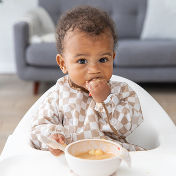 A baby in a high chair wears a checkered bib and eats puree with their hand while holding a spoon, with a bowl of food in front of them and a sofa in the background.