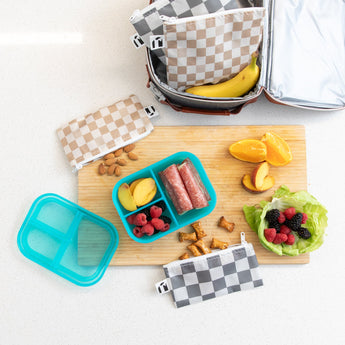 A lunch spread with a blue bento box of fruit and salami, a lettuce leaf with berries, pretzels, sliced oranges, almonds, and reusable checkered snack bags on a wooden cutting board.