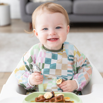A baby smiles in a Bumkins Sleeved Bib 2 Pack: Motley Club, eating waffles from a green plate while seated in a high chair.