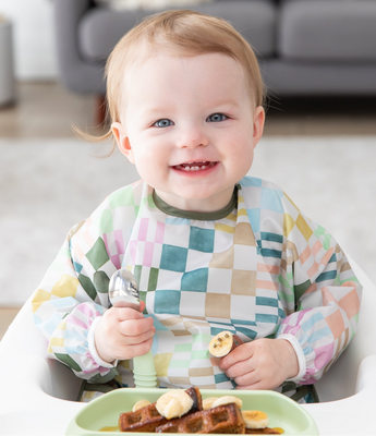 A baby smiles in a Bumkins Sleeved Bib 2 Pack: Motley Club, eating waffles from a green plate while seated in a high chair.