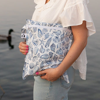 A person in a white blouse and jeans holds a bag with a blue seashell pattern near a body of water, with a duck swimming in the background.