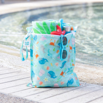 A Bumkins tan checkered wet bag filled with sunglasses and swim supplies sitting in front of a pool.