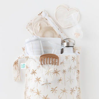 A palm tree-patterned bag holds a water bottle, notebook, hair clip, cloths, a breast pump part, and reusable rounds, all arranged neatly on a white surface.