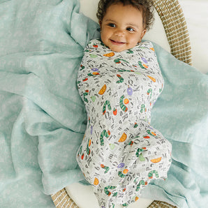 A happy baby wrapped in a LouLou Lollipop Muslin Swaddle, Eric Carle Alphabet, lying in a woven basket on a soft teal blanket.