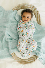 A happy baby wrapped in a LouLou Lollipop Muslin Swaddle, Eric Carle Alphabet, lying in a woven basket on a soft teal blanket.