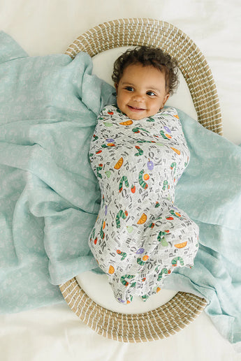 A happy baby wrapped in a LouLou Lollipop Muslin Swaddle, Eric Carle Alphabet, lying in a woven basket on a soft teal blanket.