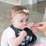Close-up of an adult hand feeding a baby wearing a black bib with a Bumkins Silicone Dipping Spoons: Minnie Mouse (Black and Pink).