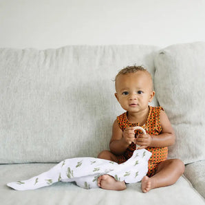 A baby in an orange romper cuddles a Bebe Au Lait Muslin Teether Blanket on a light gray couch.