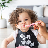 An adult hand feeds a curly-haired child wearing a black bib with a Bumkins Silicone Dipping Spoons: Minnie Mouse (Black and Pink).