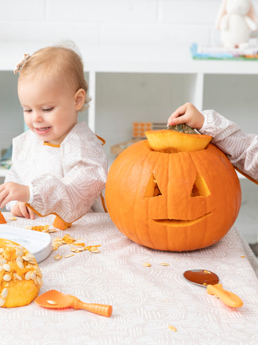 Two  girls in matching beige sleeved bibs play with a carved pumpkin on a matching tablecloth among carving spoons and pumpkin seeds.