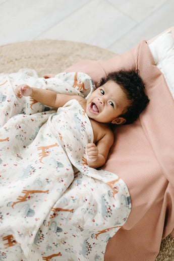 A smiling baby snuggles in the LouLou Lollipop Muslin Swaddle, Baby Dinomite, wrapped in soft bamboo muslin fabric, in a cute pink outfit.