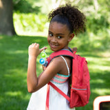 A smiling girl with curly hair holds Simpl Dimpl, Clear keychains and wears a red backpack in a sunny park. Brand: Fat Brain Toy Co.