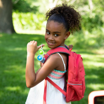 A smiling girl with curly hair holds Simpl Dimpl, Clear keychains and wears a red backpack in a sunny park. Brand: Fat Brain Toy Co.