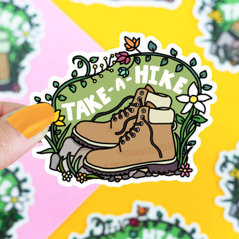 Vinyl sticker Take A Hike by Turtle Soup featuring brown hiking boots, greenery, and flowers. Held by someone with yellow nails.