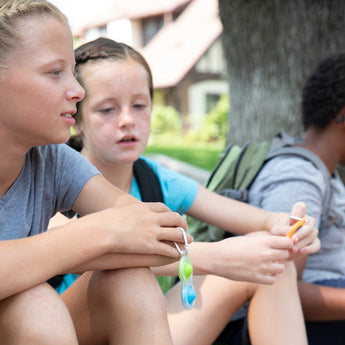 Three kids relax under a tree, two chat while one fidgets with a Fat Brain Toy Co Simpl Dimpl. Their backpacks rest beside them.