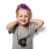 Young boy with Piggy Paints kid-safe, purple-tipped hair smiles, hands behind head, wearing a gray camera-print shirt. White background.