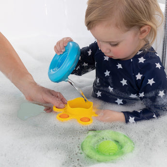 A toddler pours water using Dimpl Splash bath toys by Fat Brain Toy Co, enhancing fine motor skills in a bubble-filled bathtub.