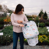 A woman in a garden holds a baby wrapped in the Milk Snob Cover Disney Enchanted Kingdoms, with plants filling the background.