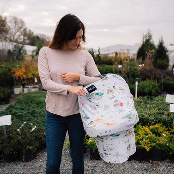A woman in a garden holds a baby wrapped in the Milk Snob Cover Disney Enchanted Kingdoms, with plants filling the background.