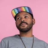 A bearded person in a Storied Hats Pride hat and gray shirt looks up against a pink backdrop. The hat reads Love is Love is Love.