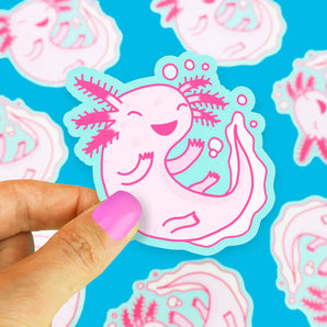 A hand holds a pink axolotl vinyl sticker by Turtle Soup against a blue background filled with identical scratch-resistant stickers.