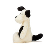 Side view of Jellycats Bashful Black & Cream Puppy Medium, a fluffy plush toy with floppy ears and a curly tail, sitting upright.