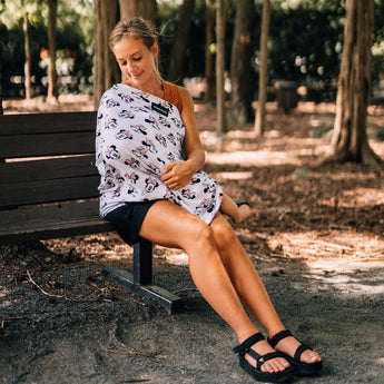 A woman sits on a bench with her baby snug in a Milk Snob Cover featuring Disneys Minnie Mouse, surrounded by trees.