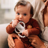 A baby in a brown outfit holds a Milk Snob Calm n Cuddle™ Cookies & Cream teether, made of food-grade silicone, sitting on an adults lap.