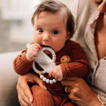 A baby in a brown outfit holds a Milk Snob Calm n Cuddle™ Cookies & Cream teether, made of food-grade silicone, sitting on an adults lap.