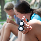 Two kids outdoors; one displays a black-and-white Fat Brain Toy Cos Simpl Dimpl fidget toy.