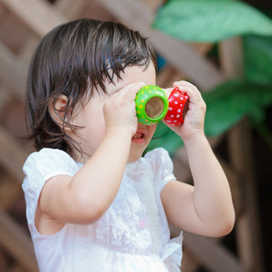 A toddler in a white dress uses PlanToys colorful cups as pretend Mushroom Kaleidoscope binoculars, surrounded by lush greenery.
