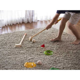 A child plays indoor mini golf with PlanToys sustainable Mini Golf Set, featuring wooden clubs and a flag hole setup on the carpet.