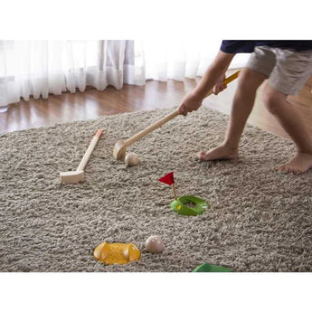 A child plays indoor mini golf with PlanToys sustainable Mini Golf Set, featuring wooden clubs and a flag hole setup on the carpet.