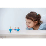 A girl in a blue shirt smiles as three PlanToys sustainably made Spinning Tops dance on the table.
