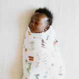 A baby sleeps peacefully wrapped in a LouLou Lollipop Cotton Swaddle Set, Farmyard, on a light-colored checkered blanket.