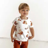 A child in a Snuggle Hunny Organic T-Shirt, Lion print, with orange pants smiles against a white background.