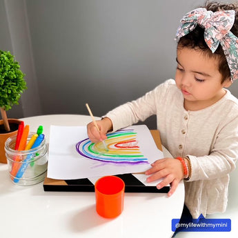 A toddler in a headband draws a vibrant rainbow at the table with OOLYs Smooth Stix Watercolor Gel Crayons; a plant peeks in.