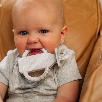 Baby on a brown chair, in a gray outfit, happily chewing on Milk Snobs Calm n Cuddle™ Teether Cookies & Cream.