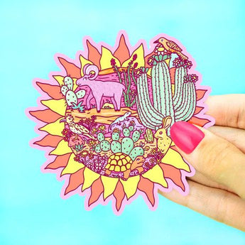 A hand displays Turtle Soups Vinyl Sticker, Desert Sun, featuring desert animals and a lizard on a bright blue background.