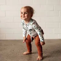 A toddler in Snuggle Hunny Organic Pants, Biscuit, and a floral top stands barefoot on concrete, smiling and leaning slightly forward.