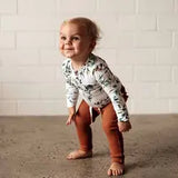 A toddler in Snuggle Hunny Organic Pants, Biscuit, and a floral top stands barefoot on concrete, smiling and leaning slightly forward.